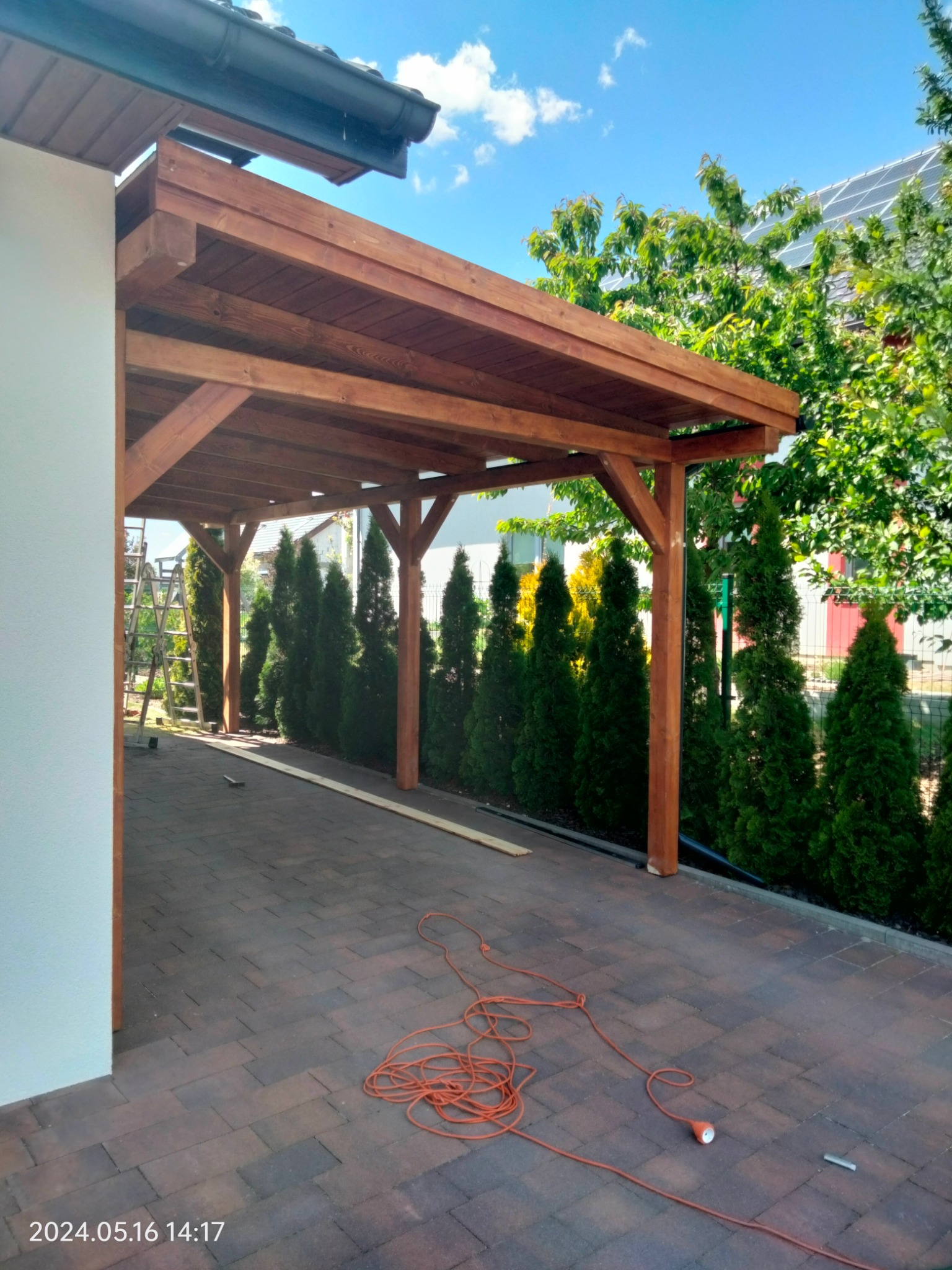 Drewniana pergola przy domu z zielonymi tujami w tle, kabel elektryczny na kostce brukowej. Widok na konstrukcję w słoneczny dzień.