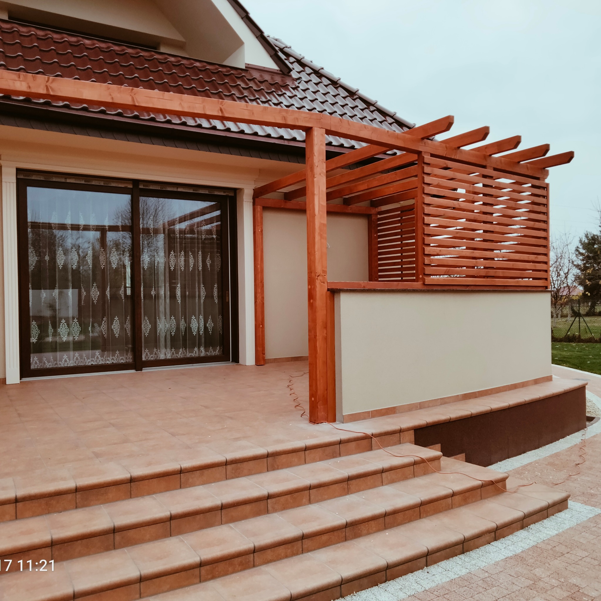 Nowoczesna pergola z drewnianymi lamelami, przylegająca do domu z tarasem wyłożonym terakotą. Widoczne schody i przeszklone drzwi przesuwne.