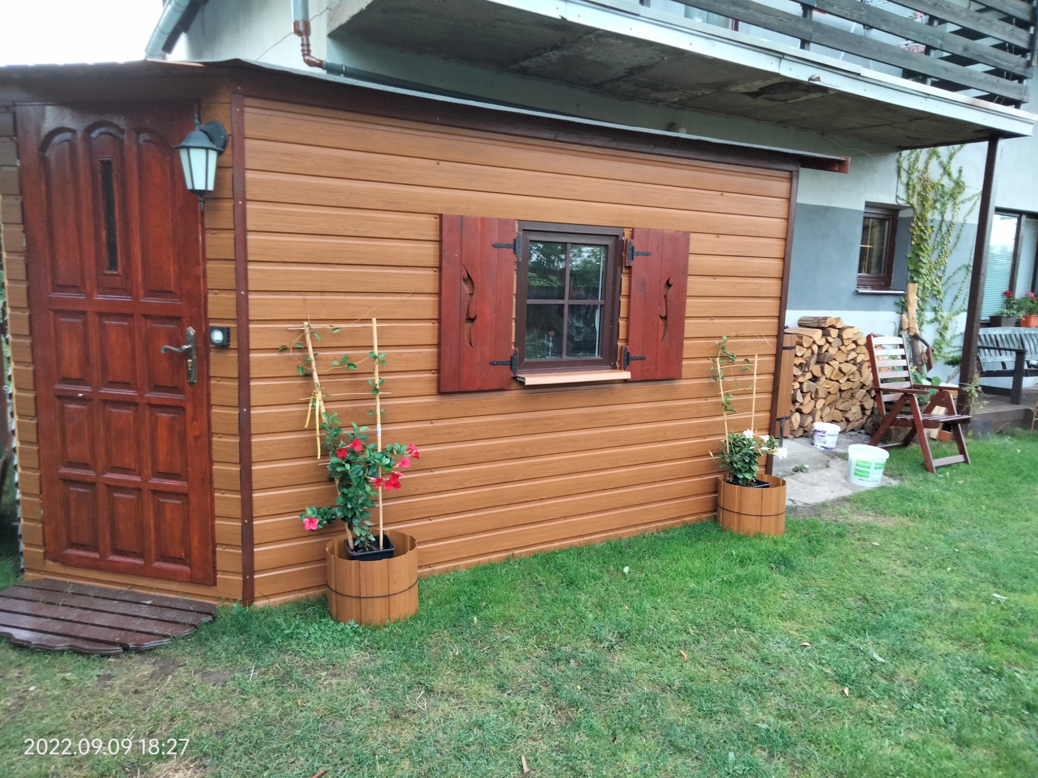 Drewniana altana z oknem i okiennicami, brązowe deski, drzwi z oświetleniem zewnętrznym, kwiaty w donicach, zielony trawnik, widok na dom z balkonem.