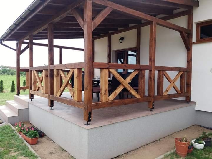 Zadaszona konstrukcja drewniana z balustradą krzyżową, widoczne schody wejściowe, donice z kwiatami. Całość w odcieniach brązu na tle jasnej elewacji budynku.