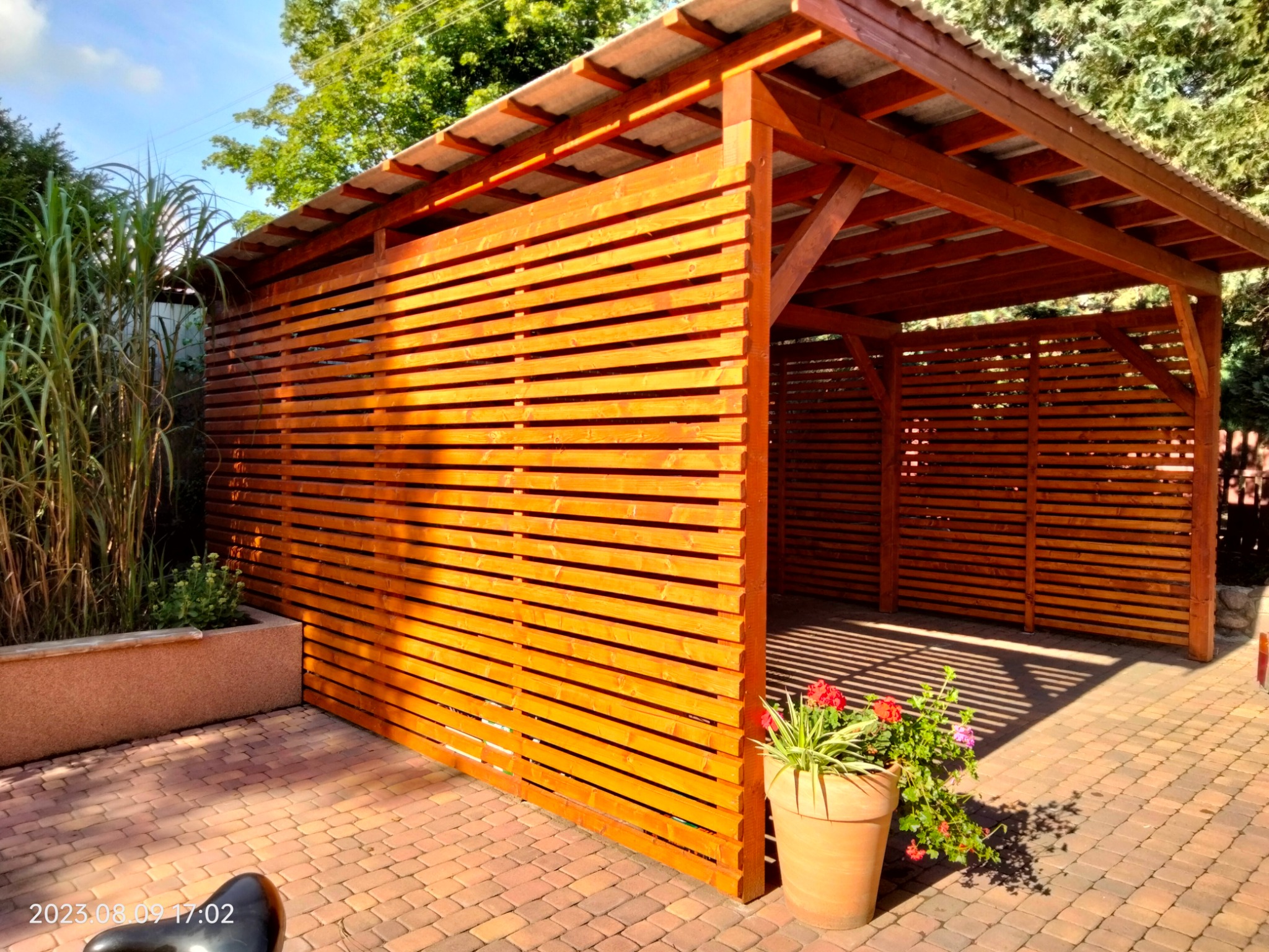 Nowoczesna, drewniana pergola z poziomymi listwami, oświetlona słońcem, z kwiatami w donicy na kostce brukowej. Widok z boku, zieleń w tle.