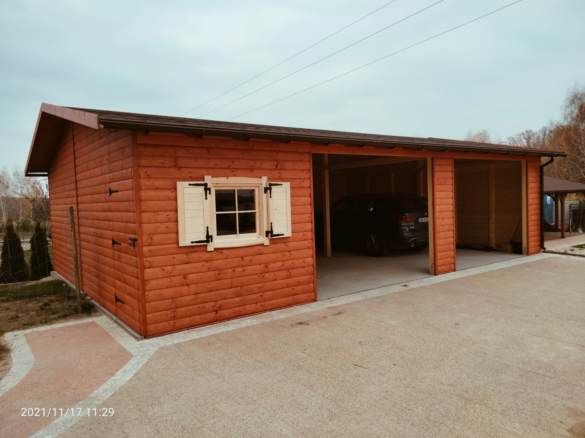 Rustykalna, drewniana konstrukcja z oknem z okiennicami i otwartymi wjazdami, w tle zaparkowany samochód. Widok z zewnątrz, na utwardzonym podłożu.