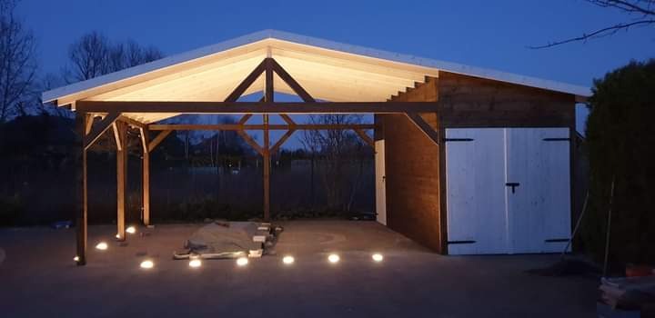 Oświetlona drewniana pergola z zadaszeniem i przylegającym budynkiem gospodarczym o zmierzchu. Podświetlenie punktowe w posadzce.