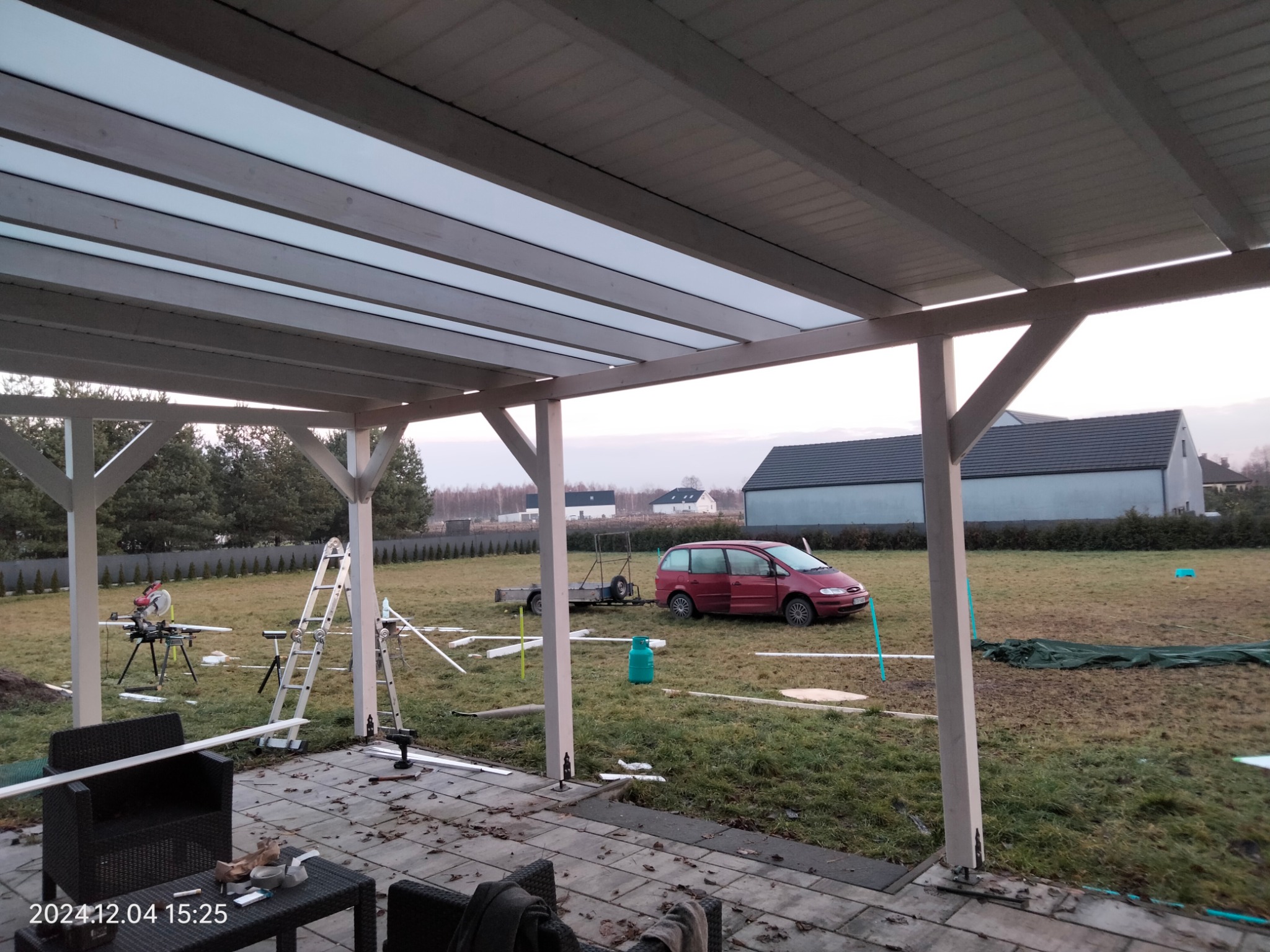 Montaż zadaszenia tarasu z przezroczystym dachem. Widoczne narzędzia, drabina i samochód w tle. Ujęcie z perspektywy tarasu.