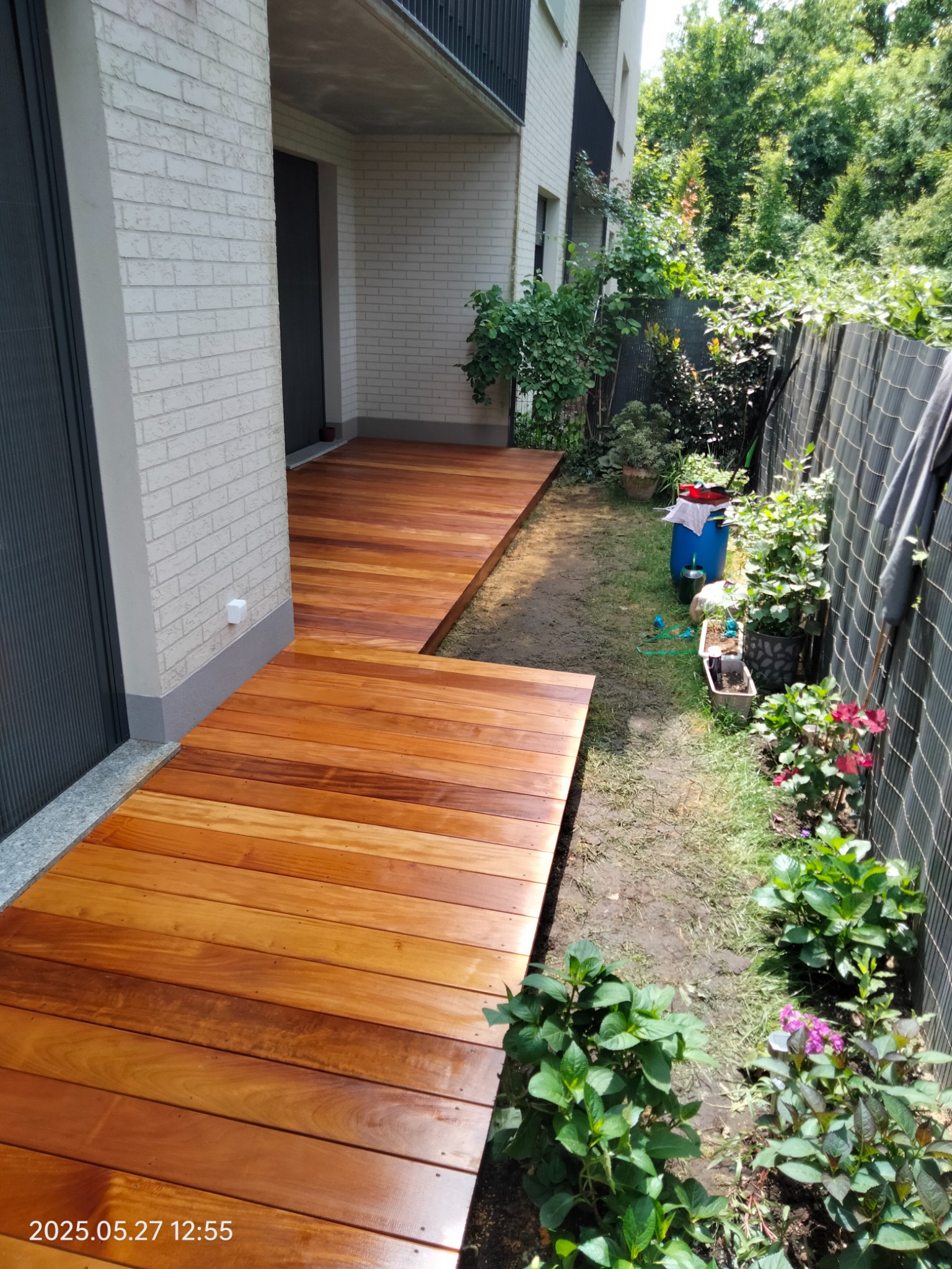 Jasnobrązowy, drewniany taras przylegający do budynku z cegły, otoczony zielenią. Widoczne deski tarasowe i fragment ogrodu z roślinnością.