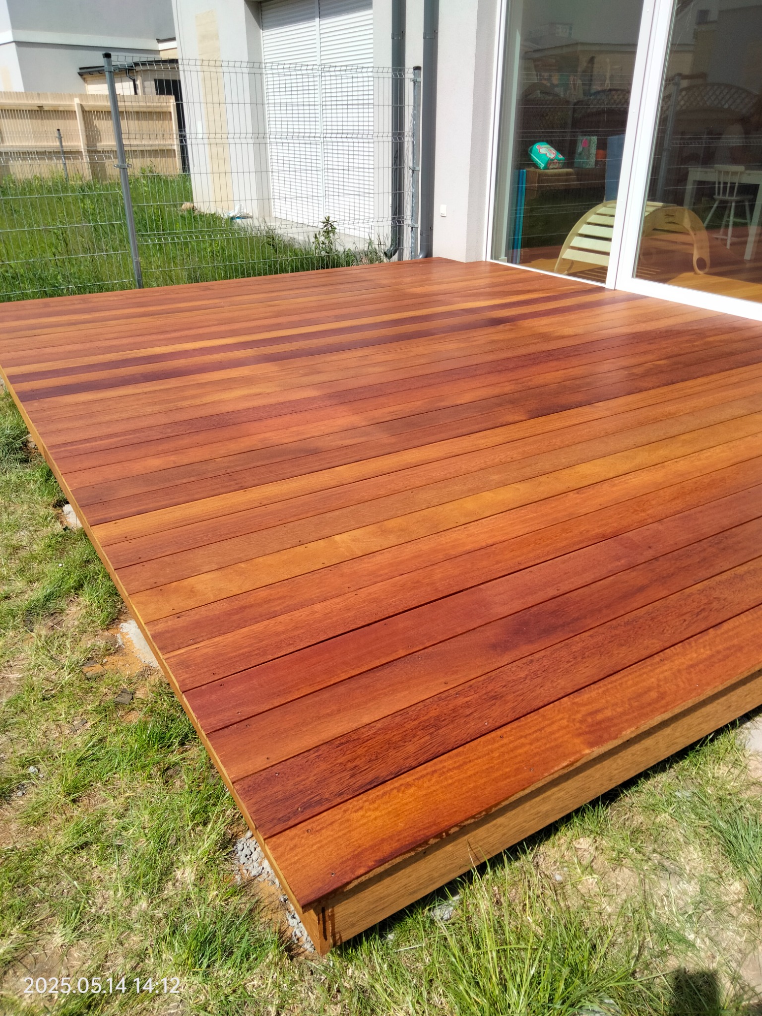 Nowy, drewniany taras z desek o ciepłej, czerwono-brązowej barwie, przylegający do budynku z dużym oknem. Widok na trawnik i ogrodzenie z siatki.