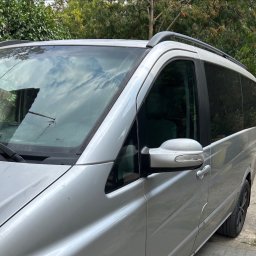 Hubertmov - Srebrny bus z relingami dachowymi, widok z boku, zarys drzew w tle odbija się w szybach. Auto zaparkowane na żwirowym podłożu.