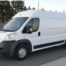 Hubertmov - Biały, wysoki furgon Peugeot na asfalcie, z widoczną tablicą rejestracyjną. Tło stanowią drzewa i betonowy mur. Dobre oświetlenie, perspektywa z boku.