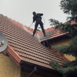 Sebastian Dudek Hyper-Tech - Pracownik w kombinezonie myje czerwoną dachówkę myjką ciśnieniową. Widok z dołu na dom z anteną satelitarną i fragmentem iglastego drzewa.