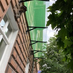 Sebastian Dudek Hyper-Tech - Elewacja budynku z dekoracyjnymi rzeźbami, zabezpieczona zieloną siatką ochronną na rusztowaniu, widoczna z dołu przez gałęzie drzew.