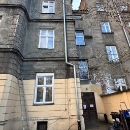 Sebastian Dudek Hyper-Tech - Stara kamienica z nową rynną, drabina oparta o ścianę. Widoczne narzędzia i fragmenty rynny na ziemi. Elewacja częściowo odnowiona, z widocznymi śladami graffiti.