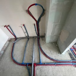 Sebastian Dudek Hyper-Tech - Rozbudowana instalacja hydrauliczna z rurami w kolorach czerwonym i niebieskim, zamocowanymi na surowym betonie podłogi i ściany. Widoczne elementy mocujące i surowy stan wykończenia.