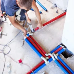 Hydraulik łączy czerwone i niebieskie rury PEX za pomocą złączek, instalacja podtynkowa na surowym podłożu, narzędzia w tle.