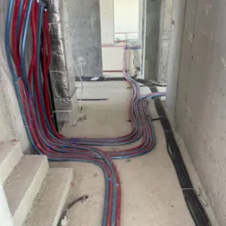 Instalacja rur grzewczych (czerwone i niebieskie) w surowym wnętrzu budynku, prowadzone wzdłuż ścian i podłogi, widoczne również rury czarne. Manometr na rurze.