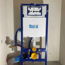 Sebastian Dudek Hyper-Tech - Stelaż podtynkowy WC Roca z niebieską ramą i podłączeniami hydraulicznymi w surowym wnętrzu łazienki, przed zabudową. Widoczne elementy instalacji.