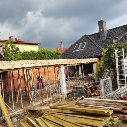 BUDOWA I REMONT Grzegorz Pietraszko - Budowa domu: widok z dołu na szalunek stropu, wsparty na stemplach, z pracownikami w tle. Na pierwszym planie deski szalunkowe. Ponure niebo w tle.