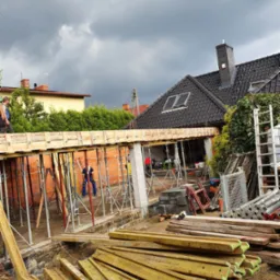 Budowa domu: widok z dołu na szalunek stropu, wsparty na stemplach, z pracownikami w tle. Na pierwszym planie deski szalunkowe. Ponure niebo w tle.