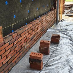 BUDOWA I REMONT Grzegorz Pietraszko - Ceglana ściana klinkierowa w trakcie budowy, widoczne ocieplenie z niebieskimi mocowaniami, ułożone cegły na folii ochronnej. Detal budowy domu.