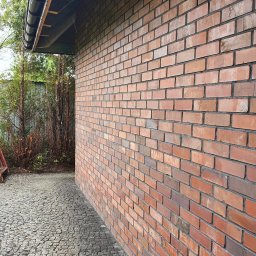 BUDOWA I REMONT Grzegorz Pietraszko - Czerwona ściana z cegły klinkierowej, widok z boku, z brukowanym chodnikiem i fragmentem dachu z rynną. Widoczne zacieki na cegłach.