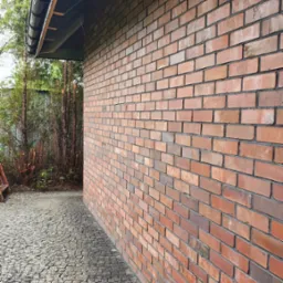 Czerwona ściana z cegły klinkierowej, widok z boku, z brukowanym chodnikiem i fragmentem dachu z rynną. Widoczne zacieki na cegłach.