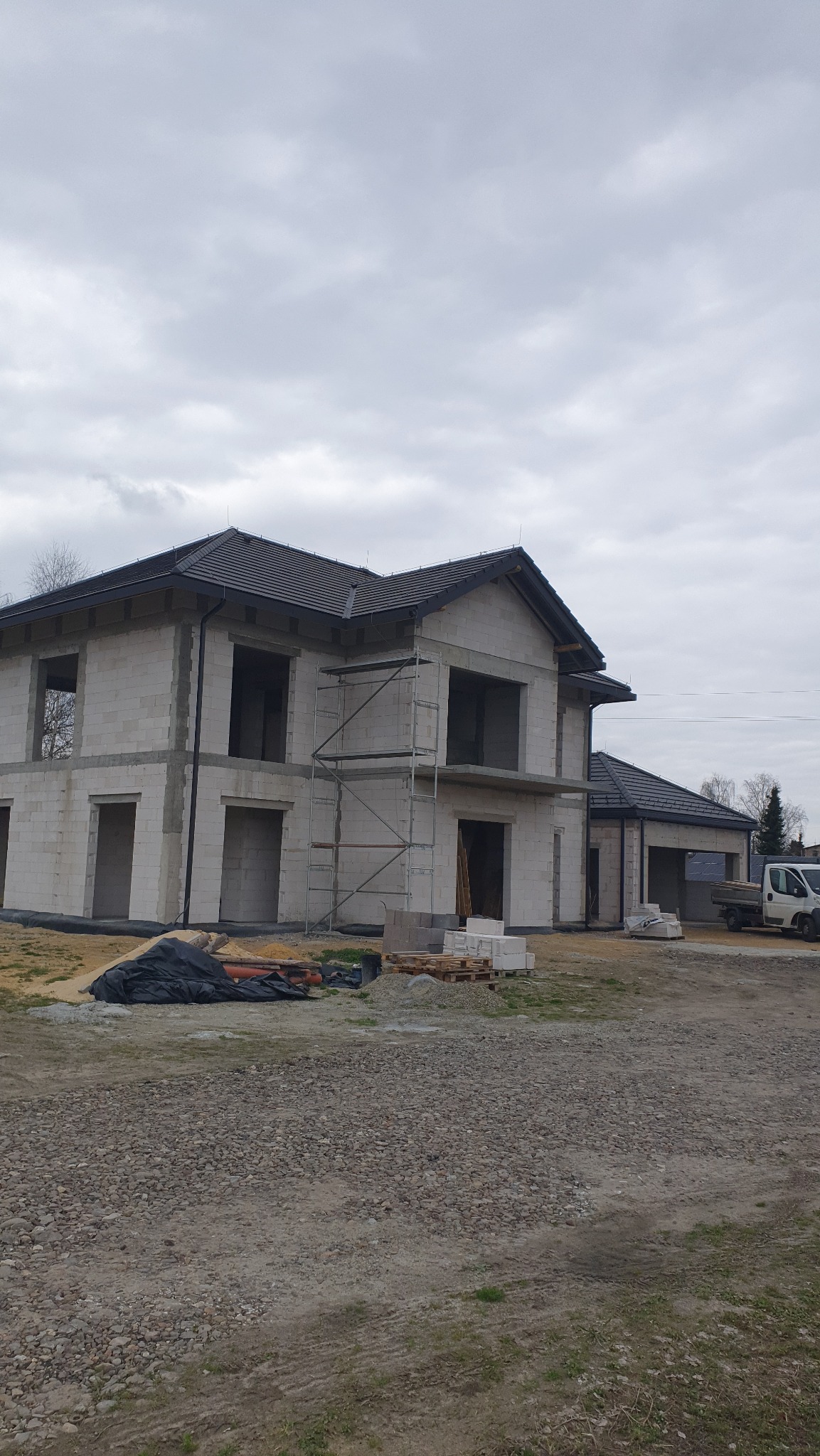 Dwukondygnacyjny dom w stanie surowym otwartym, z szarym dachem i rusztowaniem. Widoczne palety z bloczkami i gruz. Pochmurne niebo w tle.