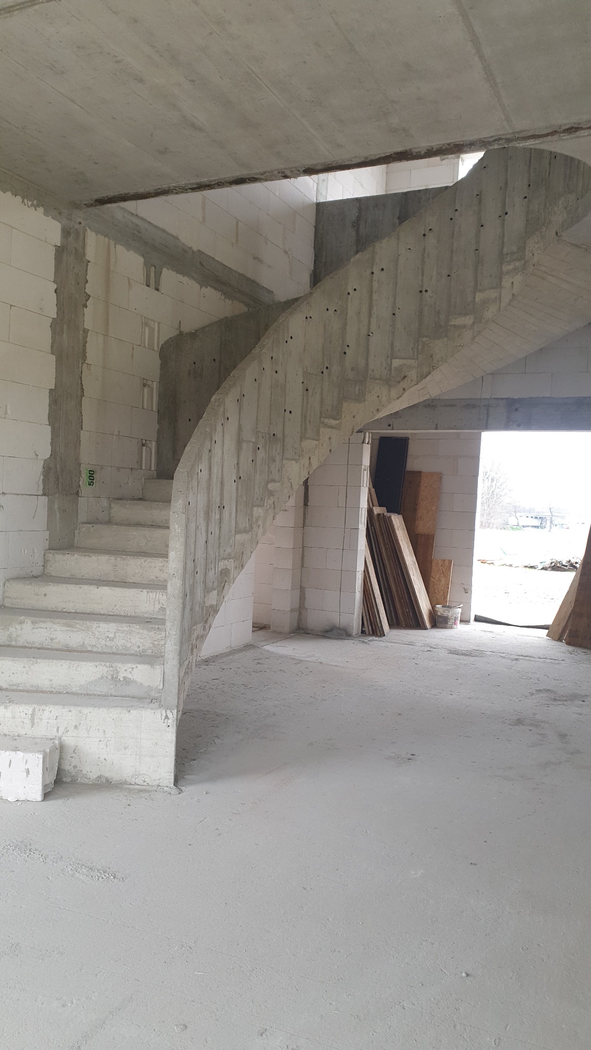 Surowe schody spiralne z betonu wewnątrz budynku w trakcie budowy, widoczne białe bloczki i materiały budowlane. W tle jasne światło dzienne.
