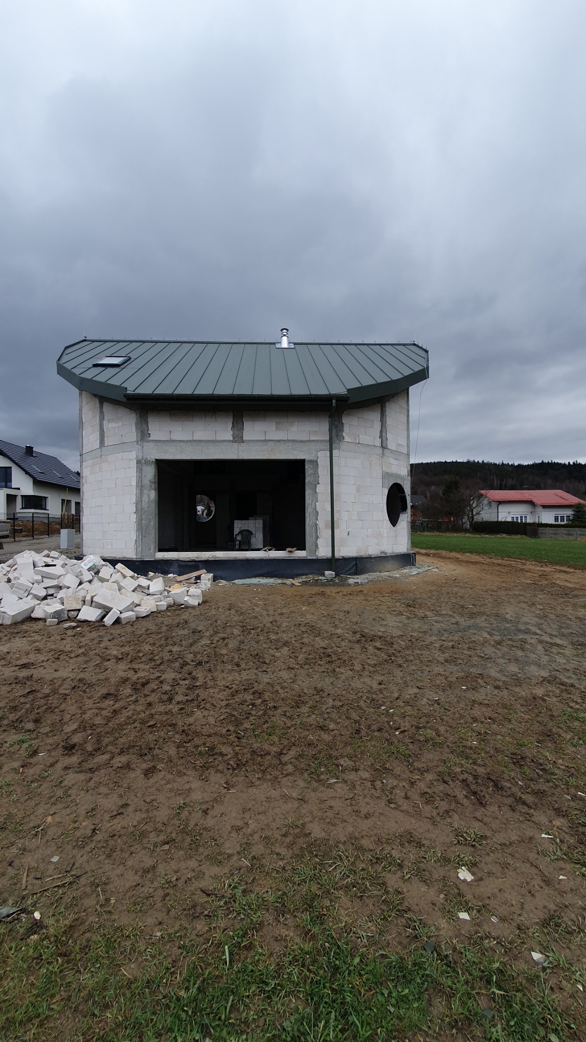 Niedokończony, okrągły dom z białych bloczków, z szarym dachem i stertą materiałów budowlanych na błotnistym podłożu, w tle zabudowa jednorodzinna.