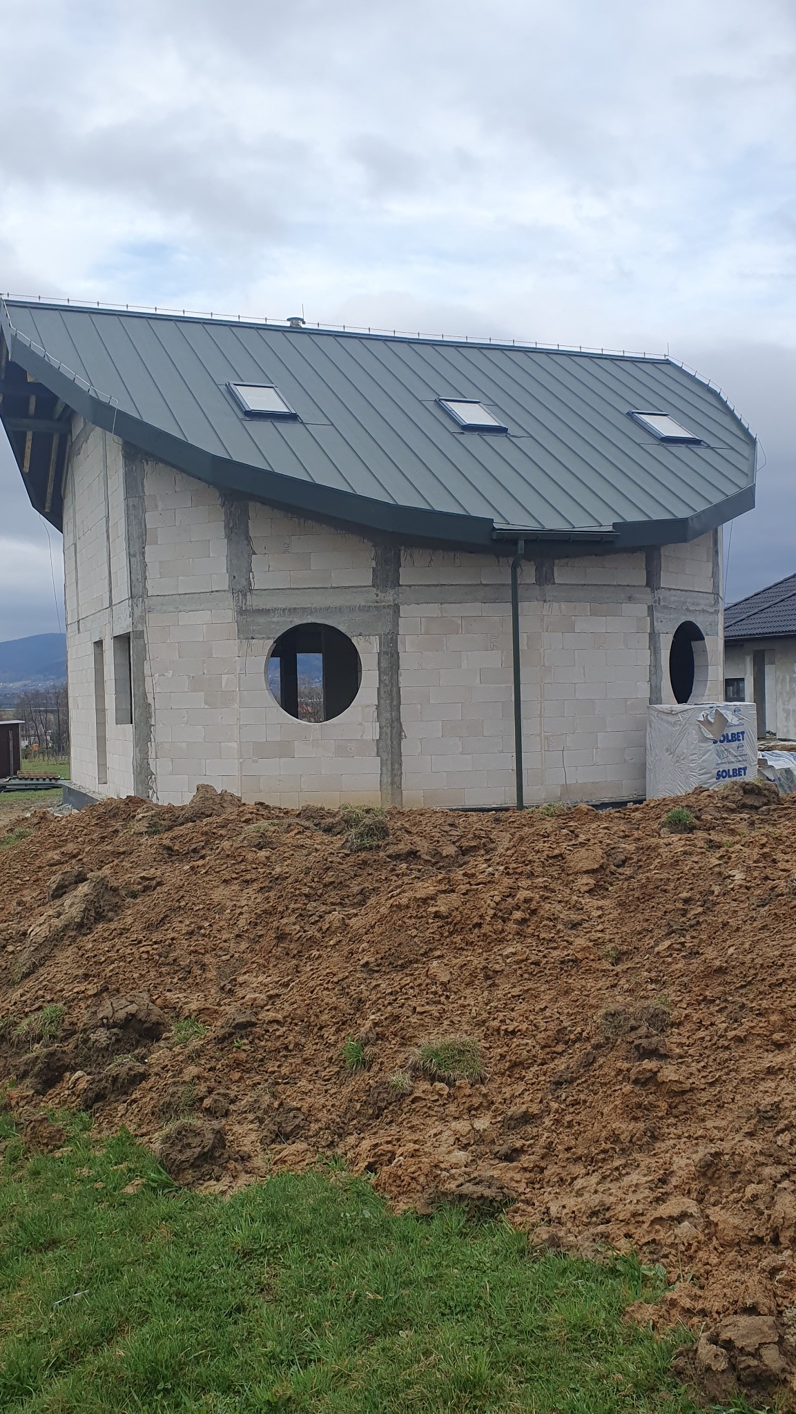 Niedokończony dom z betonu komórkowego z nietypowym dachem i okrągłymi oknami, na tle wzgórz i pochmurnego nieba. Widoczne bloczki SOLBET.