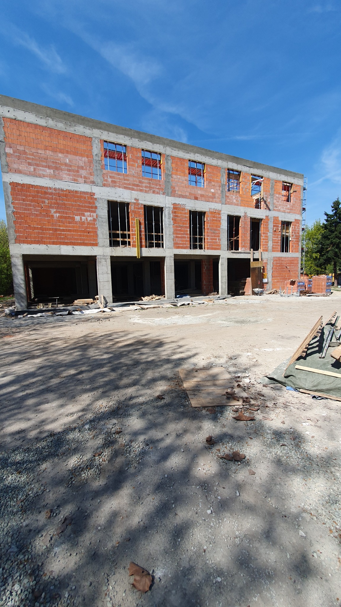 Trzykondygnacyjny budynek w stanie surowym otwartym, z czerwonej cegły i betonu, z widocznymi otworami okiennymi i wejściowymi. Błękitne niebo w tle.
