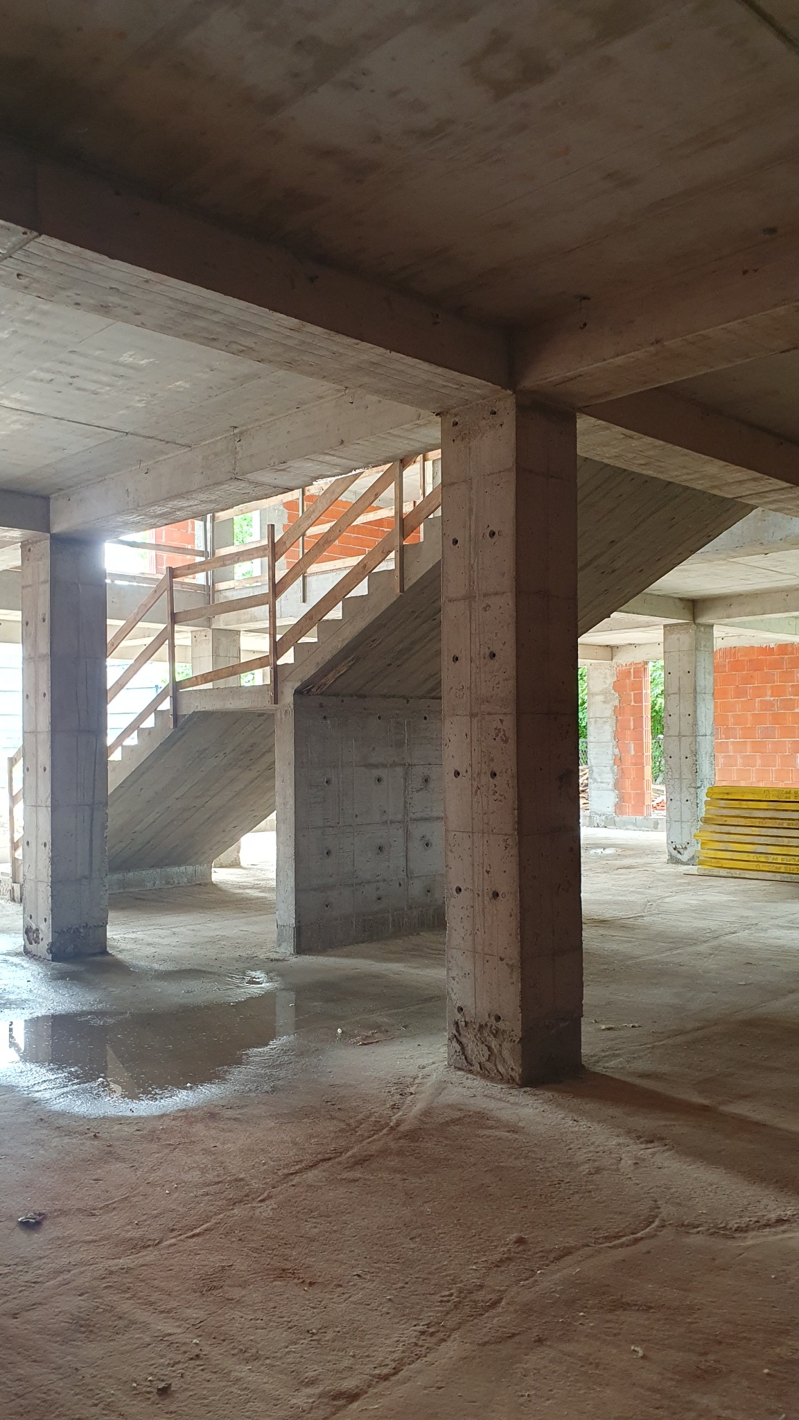 Surowy stan budynku żelbetowego w trakcie budowy, widoczne betonowe kolumny, schody z drewnianą barierką i częściowo wymurowane ściany. Na podłodze woda.