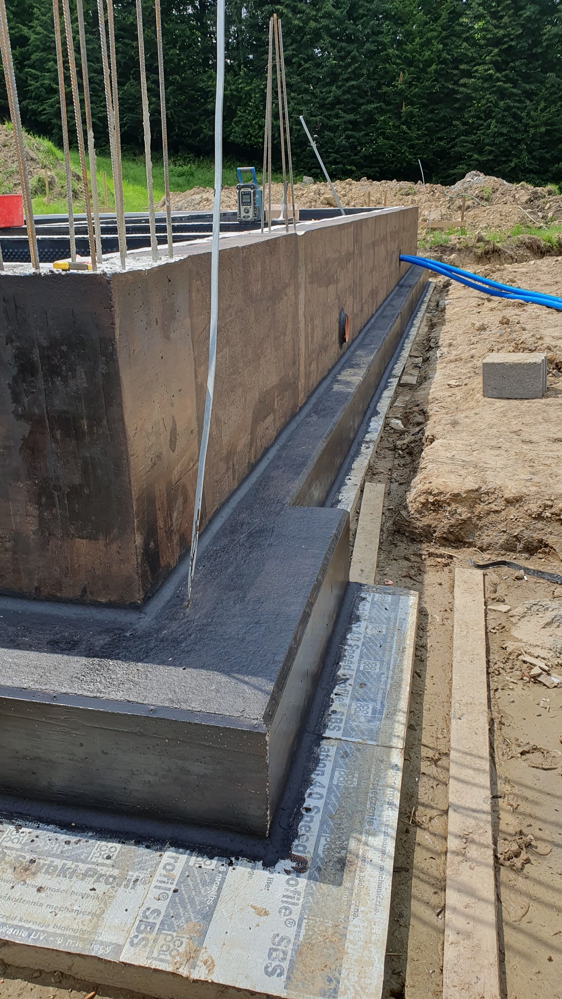 Zabetonowany fundament domu z wystającymi prętami zbrojeniowymi, izolacją przeciwwilgociową i niebieskimi rurami w ziemi. Widoczne narzędzia i poziomica.