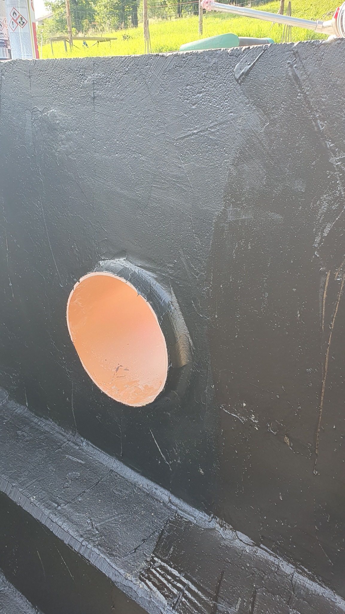 Hydroizolacja fundamentu z rurą kanalizacyjną, pokryta czarną masą bitumiczną. Widoczne ślady pędzla i nierówności powierzchni. Zabezpieczenie przed wilgocią.