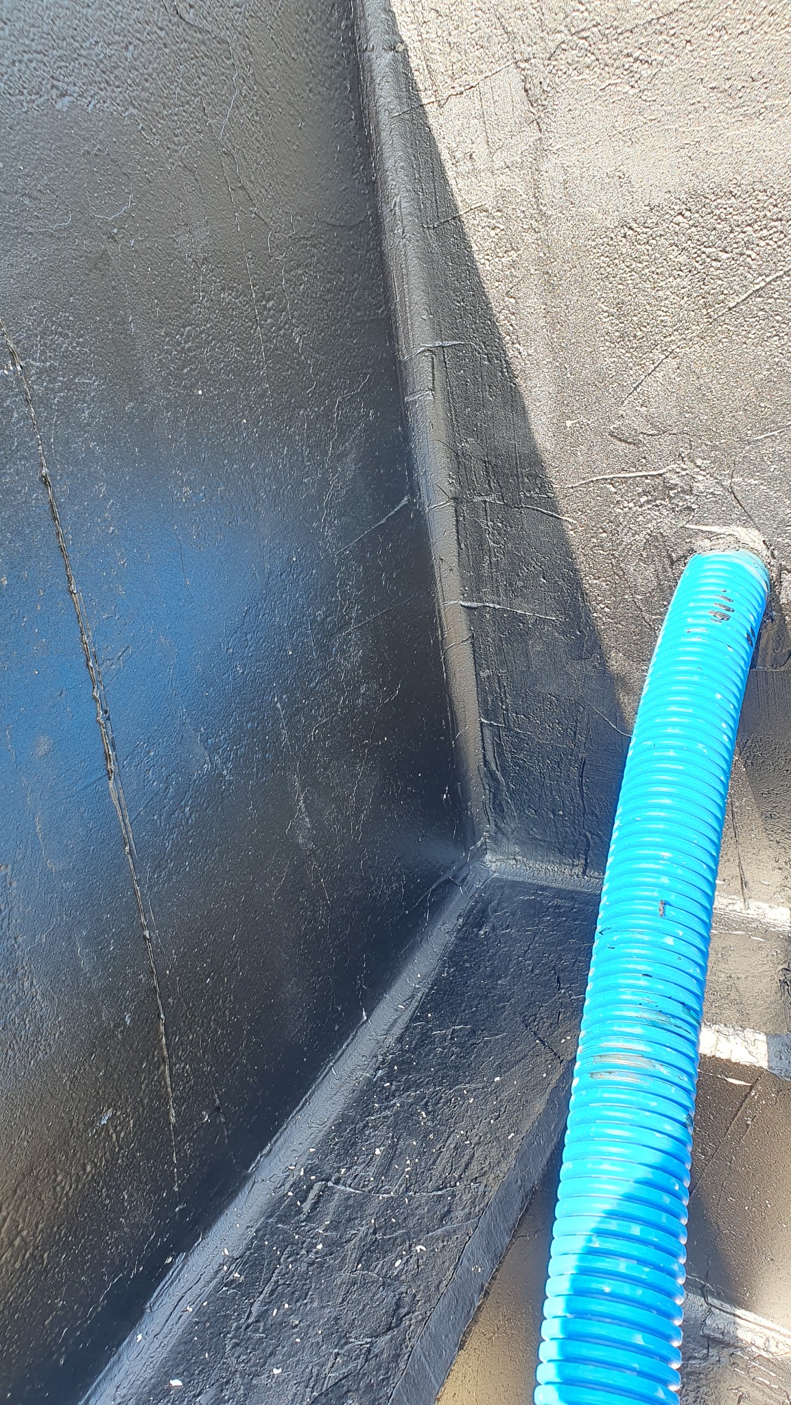 Hydroizolacja fundamentu z niebieską rurą drenażową. Czarna, błyszcząca powłoka izolacyjna na szarej ścianie fundamentowej. Detal prac budowlanych.