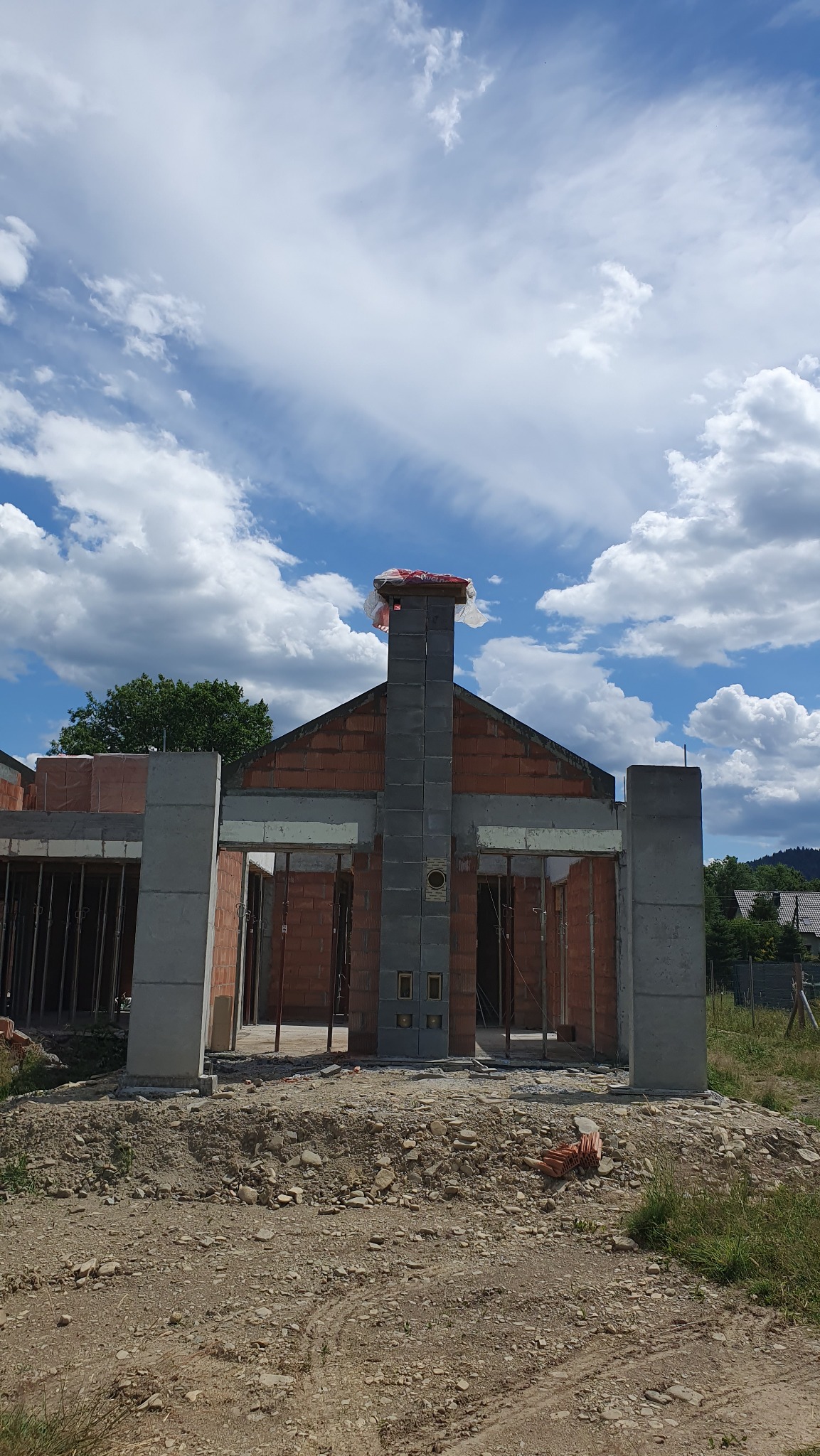 Wznoszenie domu z czerwonej cegły i szarego betonu w słoneczny dzień. Widoczny komin z szarych pustaków. Niebo błękitne z białymi chmurami. Surowy stan budynku.