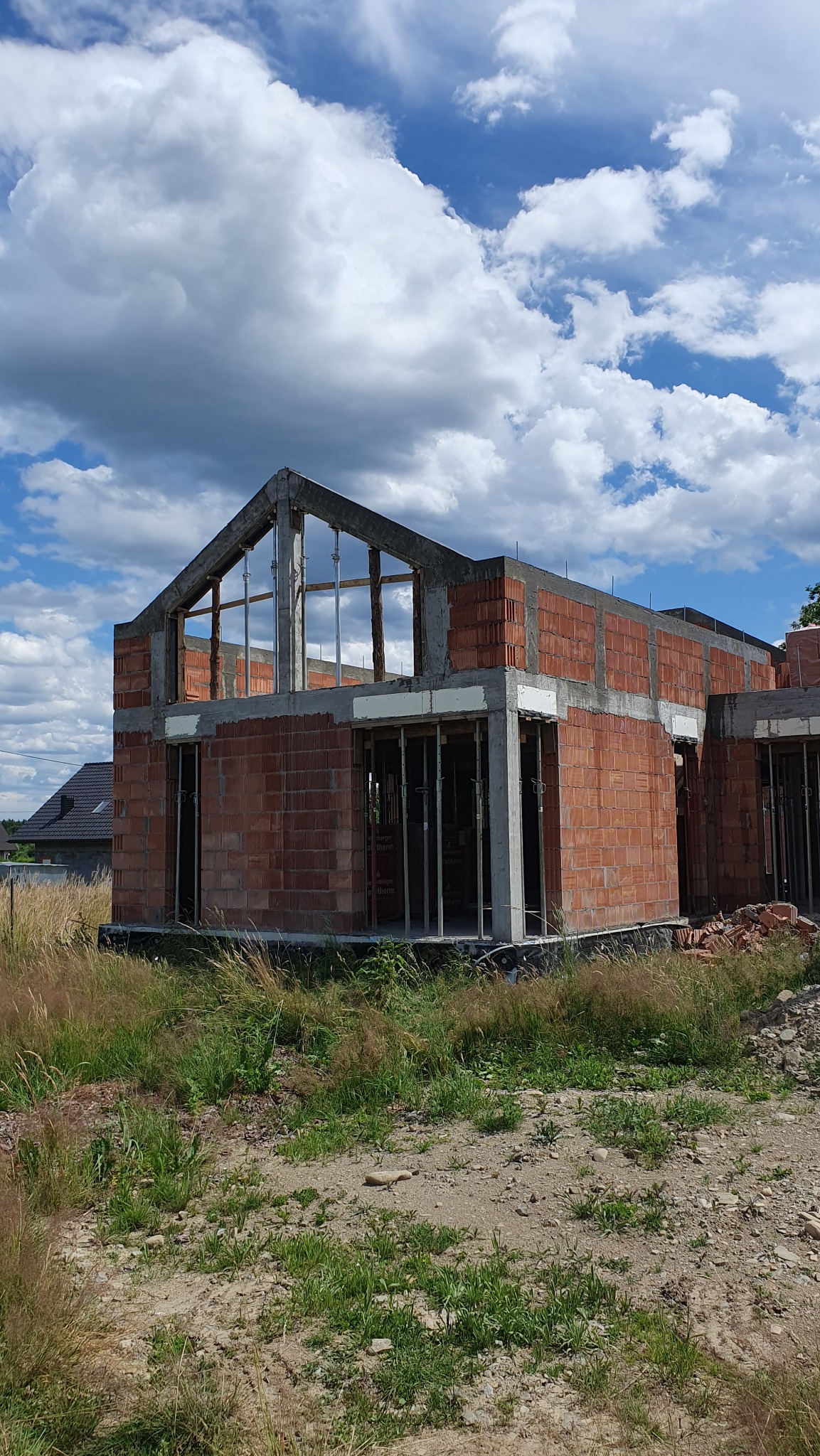 Budowa domu z czerwonej cegły w Lipowej, w stanie surowym otwartym. Widoczne ściany i betonowe elementy konstrukcyjne, na tle błękitnego nieba z chmurami.