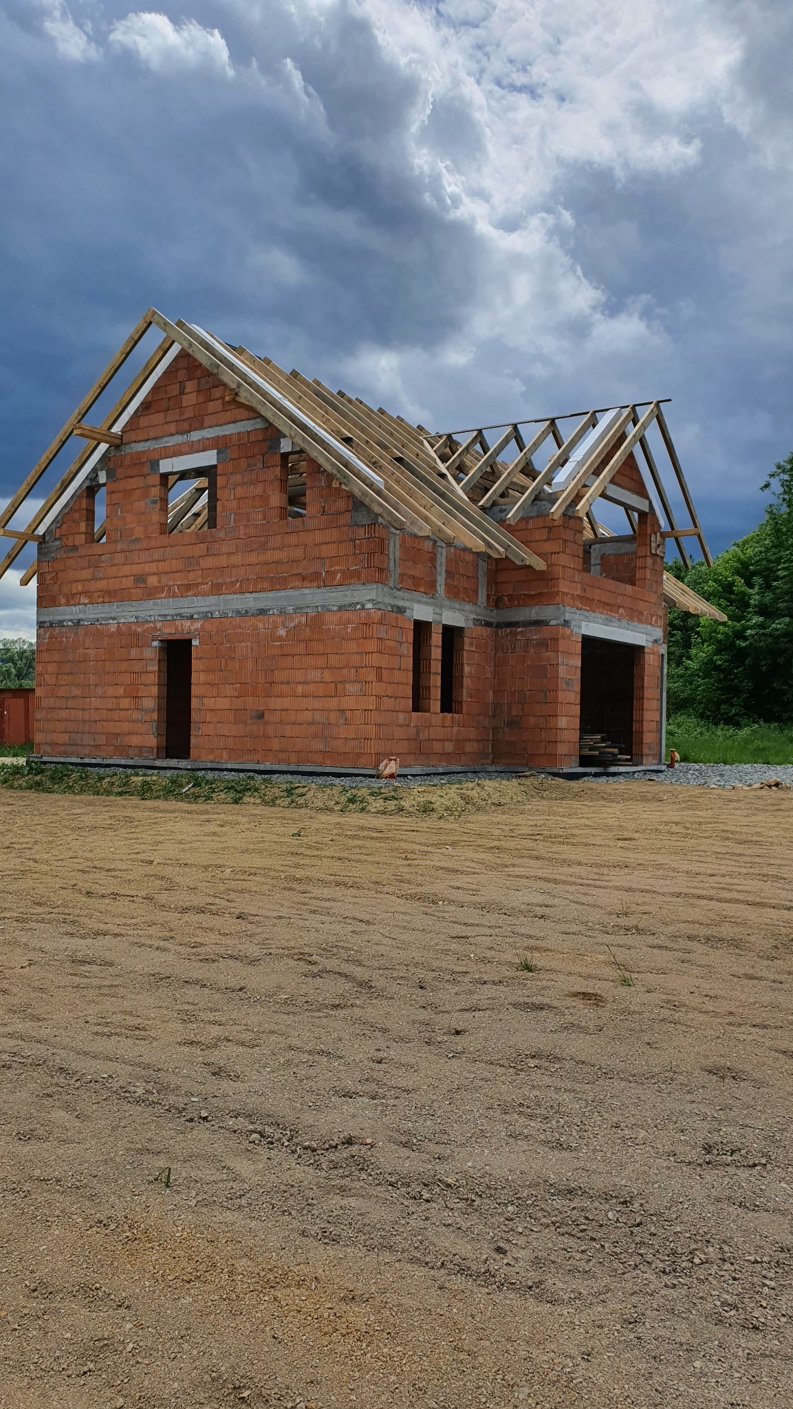 Surowy stan domu z czerwonej cegły z drewnianą konstrukcją dachu na tle pochmurnego nieba. Widoczne otwory okienne i drzwiowe.