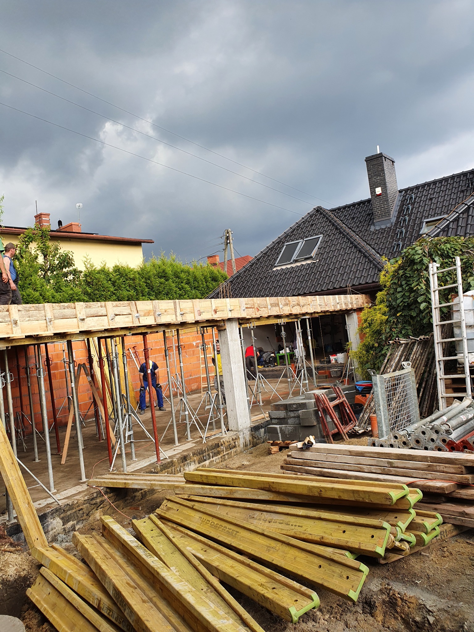 Budowa domu: widok z dołu na szalunek stropu, wsparty na stemplach, z pracownikami w tle. Na pierwszym planie deski szalunkowe. Ponure niebo w tle.