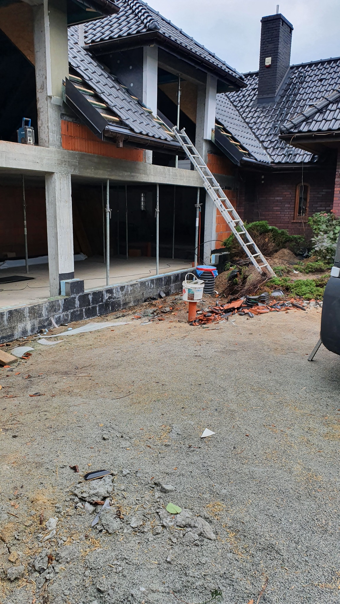 Budowa domu w trakcie realizacji: surowy stan z widocznymi elementami konstrukcyjnymi, rusztowaniami i materiałami budowlanymi. Uwagę przyciąga drabina oparta o budynek.