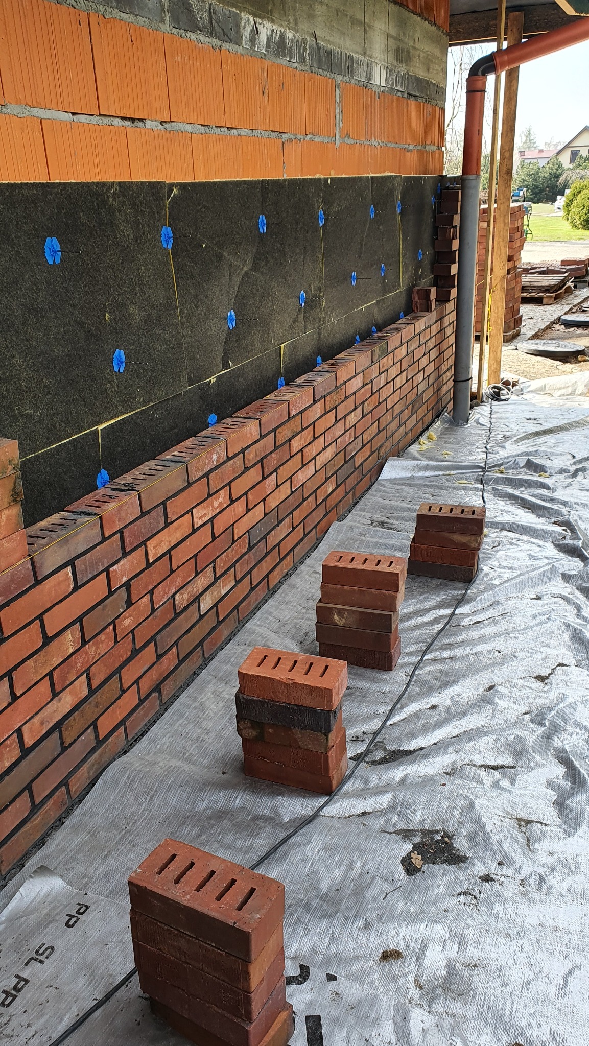 Ceglana ściana klinkierowa w trakcie budowy, widoczne ocieplenie z niebieskimi mocowaniami, ułożone cegły na folii ochronnej. Detal budowy domu.