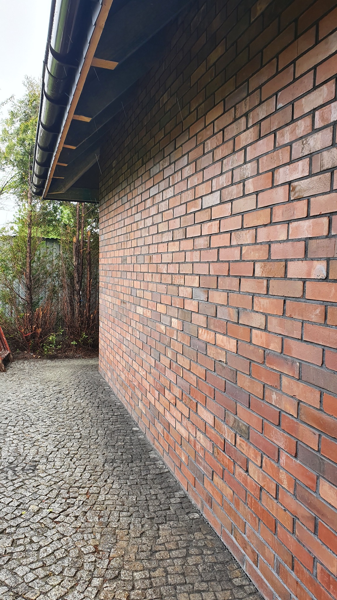 Czerwona ściana z cegły klinkierowej, widok z boku, z brukowanym chodnikiem i fragmentem dachu z rynną. Widoczne zacieki na cegłach.