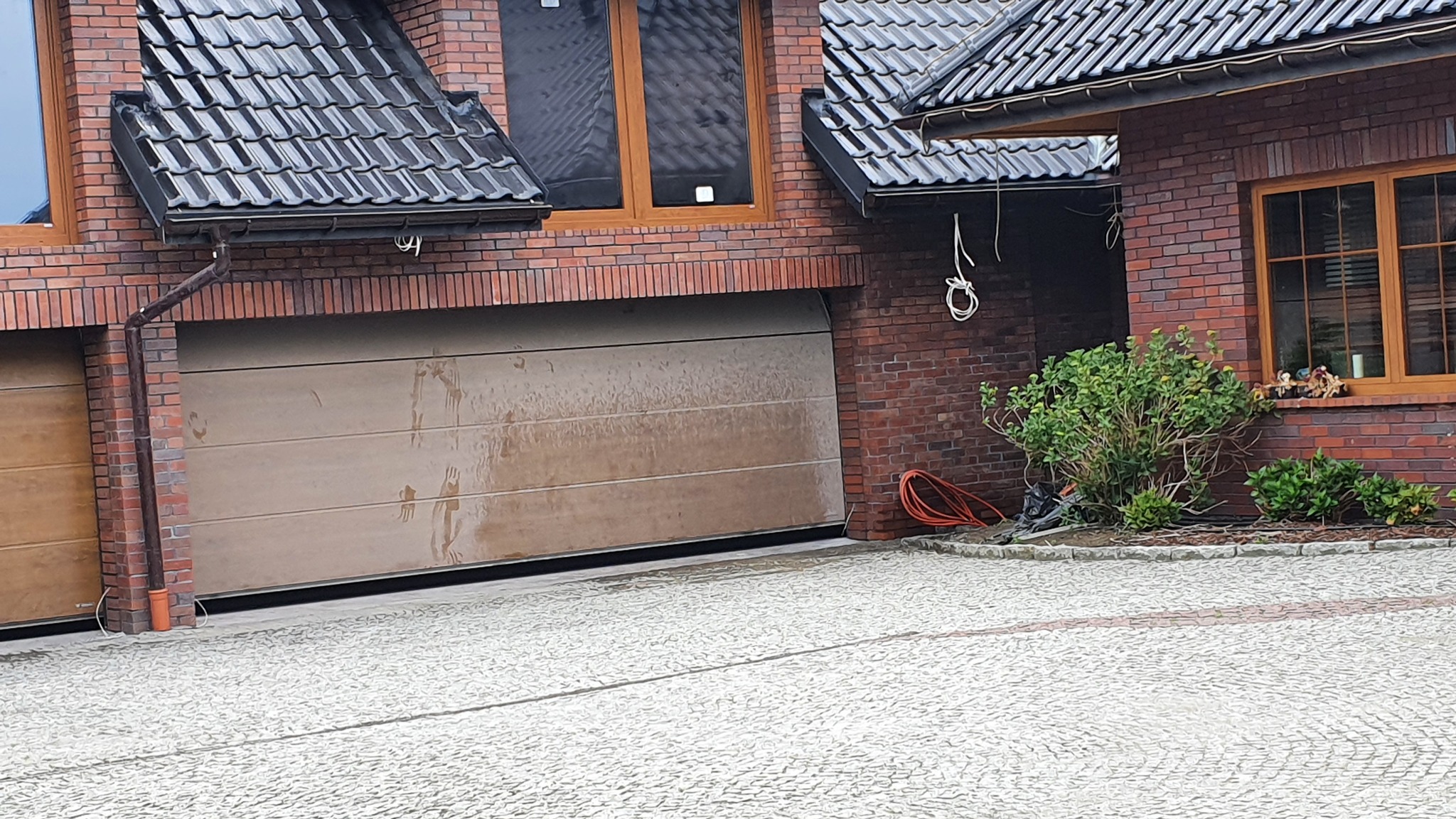 Ceglany dom z garażem: brązowa brama segmentowa z widocznymi śladami wody. Czarna dachówka i okna z drewnianą stolarką. Na zewnątrz kostka brukowa i zieleń.
