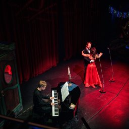 Władysław Turański - Para tańczy na scenie przy akompaniamencie pianisty. Czerwone światła, dekoracje świąteczne i ozdobna garderoba tworzą elegancką atmosferę wydarzenia.