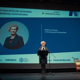 Władysław Turański - Prelegentka na scenie podczas VII Małopolskiego Kongresu Srebrnej Gospodarki. W tle ekran z logiem i zdjęciem prelegentki Elżbiety A.