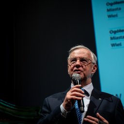 Władysław Turański - Mężczyzna w garniturze przemawia do mikrofonu na tle ekranu projekcyjnego z tekstem. Ujęcie z dołu, widoczny fragment dekoracji scenicznej.