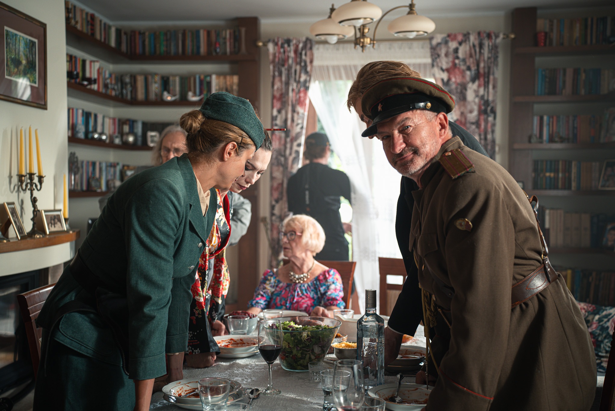 Scena z planu filmowego: aktorzy w kostiumach (lata 40.) stoją przy stole z jedzeniem. W tle reżyser i ekipa. Biblioteczka, lampa i okno w tle.
