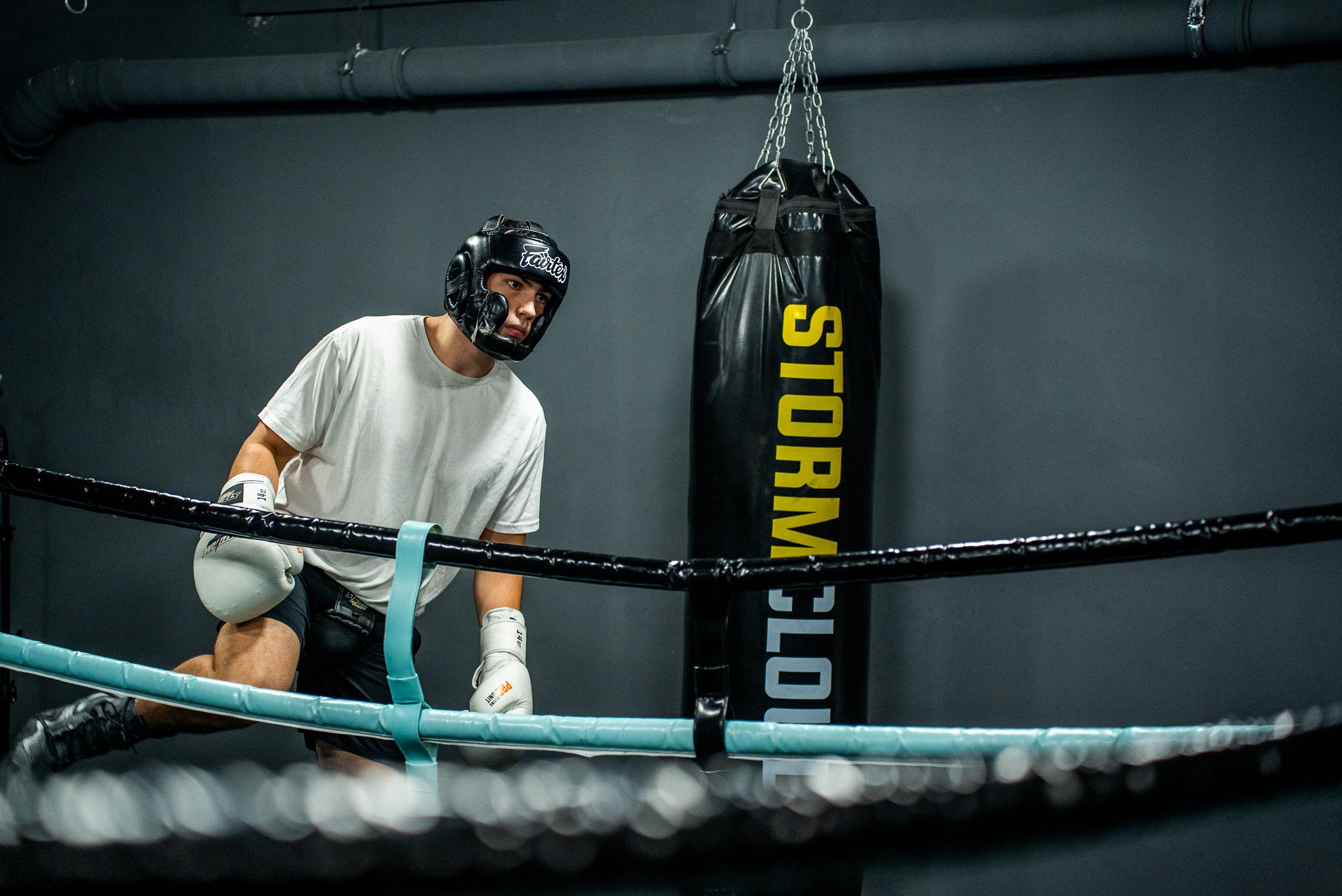 Bokser w ringu, w kasku i rękawicach, obok worka treningowego. Ujęcie z dołu, widoczne liny ringu i fragment instalacji na suficie. Ciemna kolorystyka.