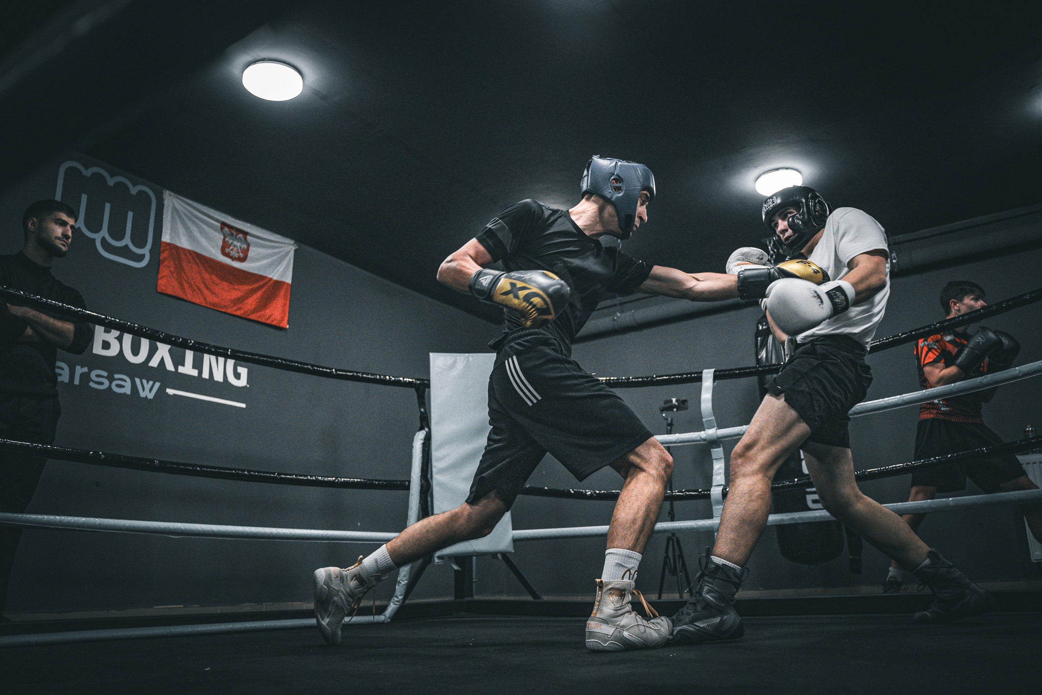 Dynamiczne ujęcie sparingu bokserskiego w ringu: bokserzy w kaskach i rękawicach, flaga Polski w tle, oświetlenie halogenowe. Intensywny trening w klubie bokserskim.
