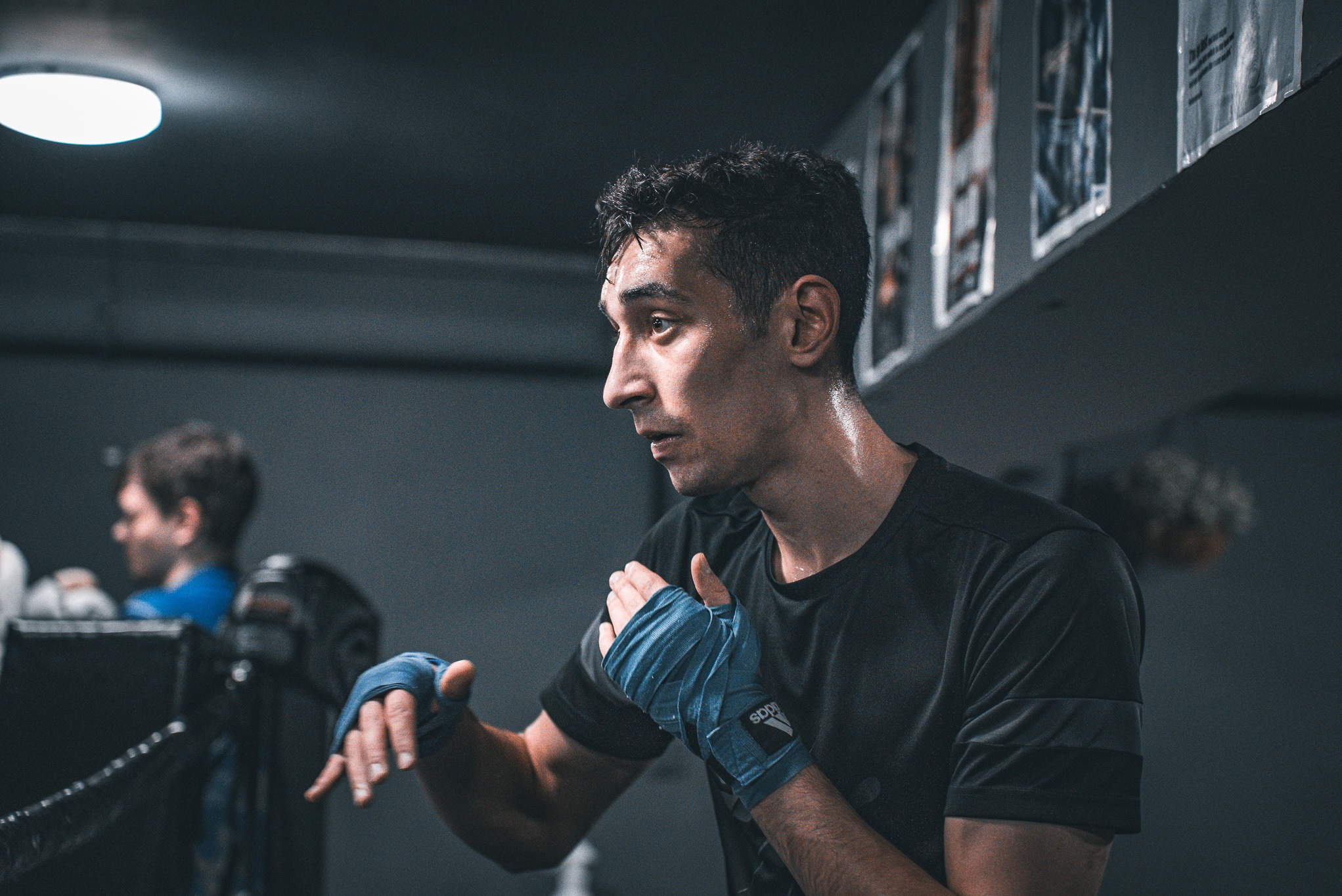Mężczyzna w ciemnej koszulce, z niebieskimi bandażami na dłoniach, przygotowuje się do treningu bokserskiego. Widoczny pot na czole, w tle ring i inne osoby.