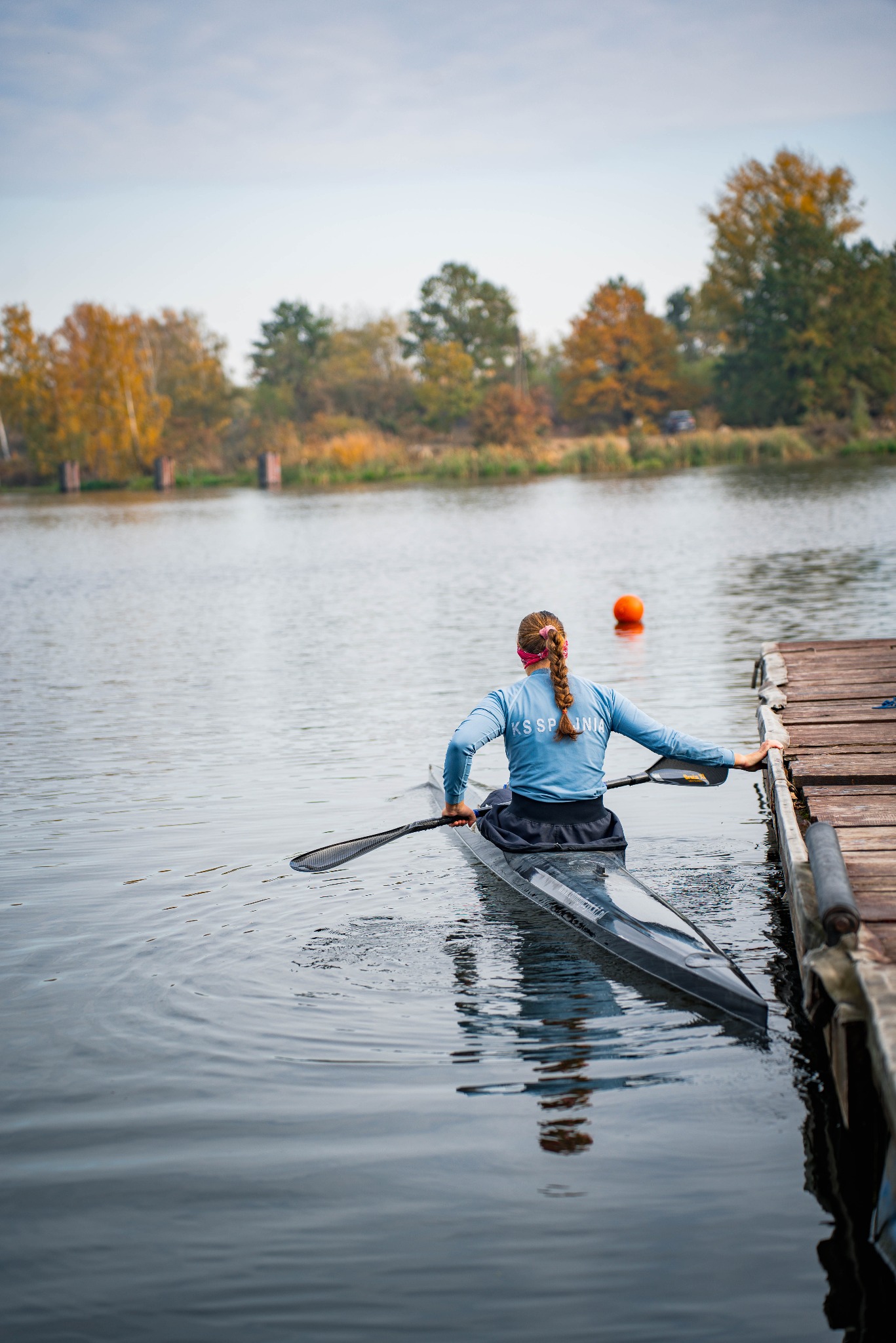 Kobieta w kajaku, widziana od tyłu, trzymająca wiosło, podpływa do drewnianego pomostu na tle jesiennego krajobrazu i spokojnej wody.