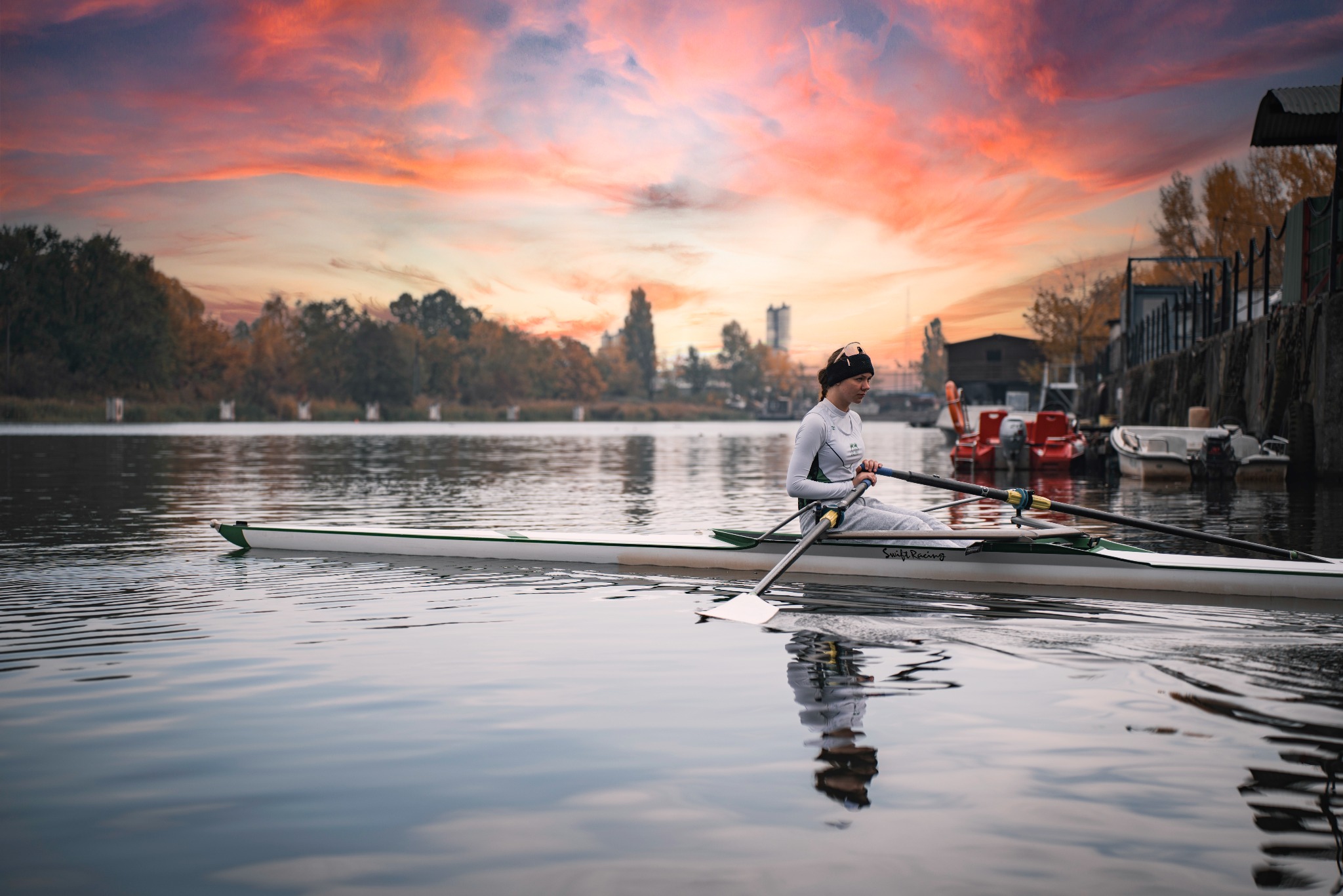 Wioślarka w łodzi na spokojnej wodzie o zmierzchu. Widoczne odbicie w wodzie, drzewa na brzegu i intensywne kolory zachodzącego słońca na niebie.